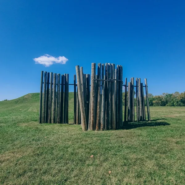 Circle of tall wooden posts representing part of Cahokia’s ancient Woodhenge solar calendar, with grassy mounds visible in the background.