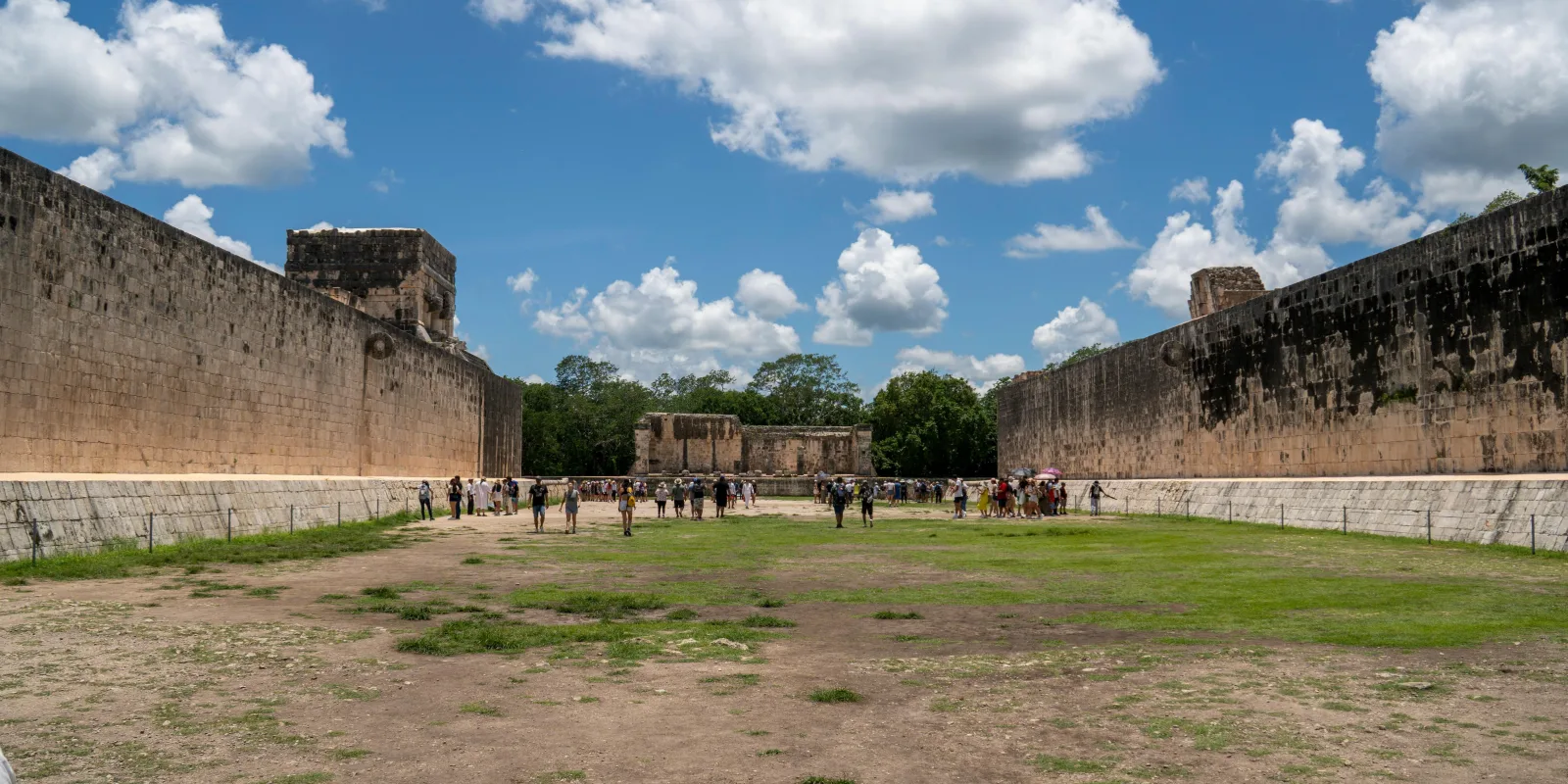 Wide view of the Great Ballcourt at Chichén Itzá with visitors walking between towering stone walls and ancient goal rings under a bright sky