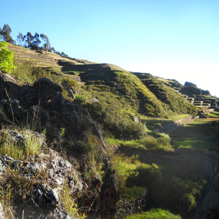 Panoramic hillside view of Chinchero showing stone terraces along the slope, natural rock formations, and vegetation under a clear blue sky.