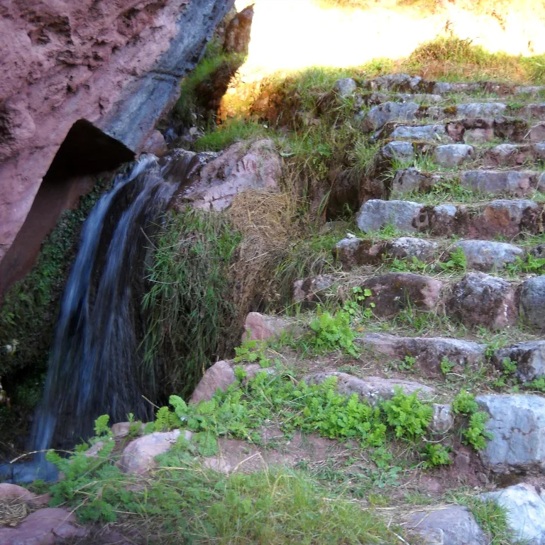 Small cascade of water emerging from a stone Inca canal beside a worn stone stairway at Chinchero, surrounded by green vegetation and pink rock.