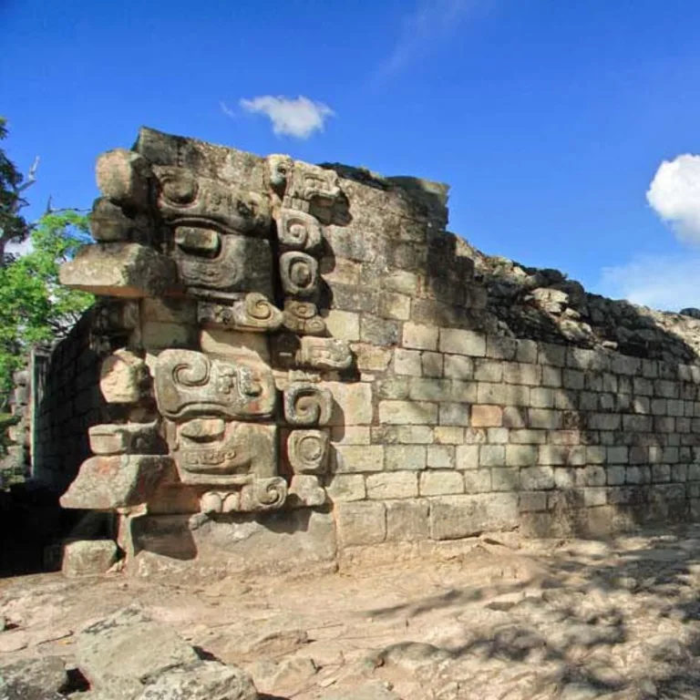 Stacked Witz mountain-monster masks on a corner of Temple 22 at Copán, Honduras, with projecting snouts, carved scrolls, and adjoining stone walls beneath a blue sky.