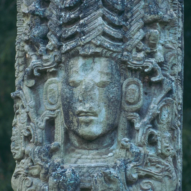 Close-up of Maya ruler Uaxaclajuun Ub’aah K’awiil, known as 18 Rabbit, on Stela A at Copán, Honduras, showing his carved face, layered headdress, large earspools, and surrounding supernatural figures.