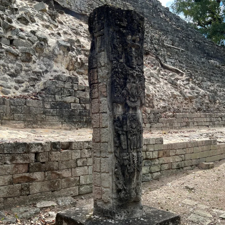 Stela N at Copán, Honduras, showing a standing Maya ruler in elaborate regalia, columns of hieroglyphs along its side, and the stone terraces of Temple 11 behind it.