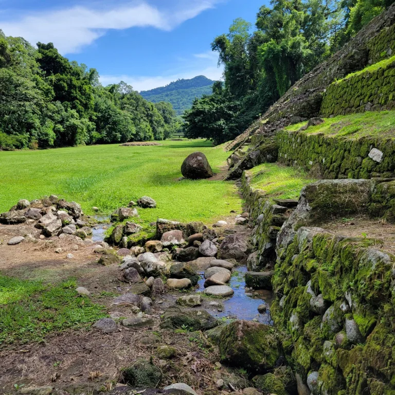 Spring-fed stream flowing through rocks and ruins at El Cuajilote, with pyramid mounds and mountains in the background.