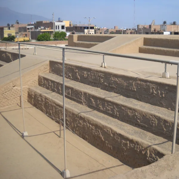Close-up of carved adobe friezes on the stepped ramp of Huaca Arco Iris (El Dragón), with Trujillo cityscape in the background