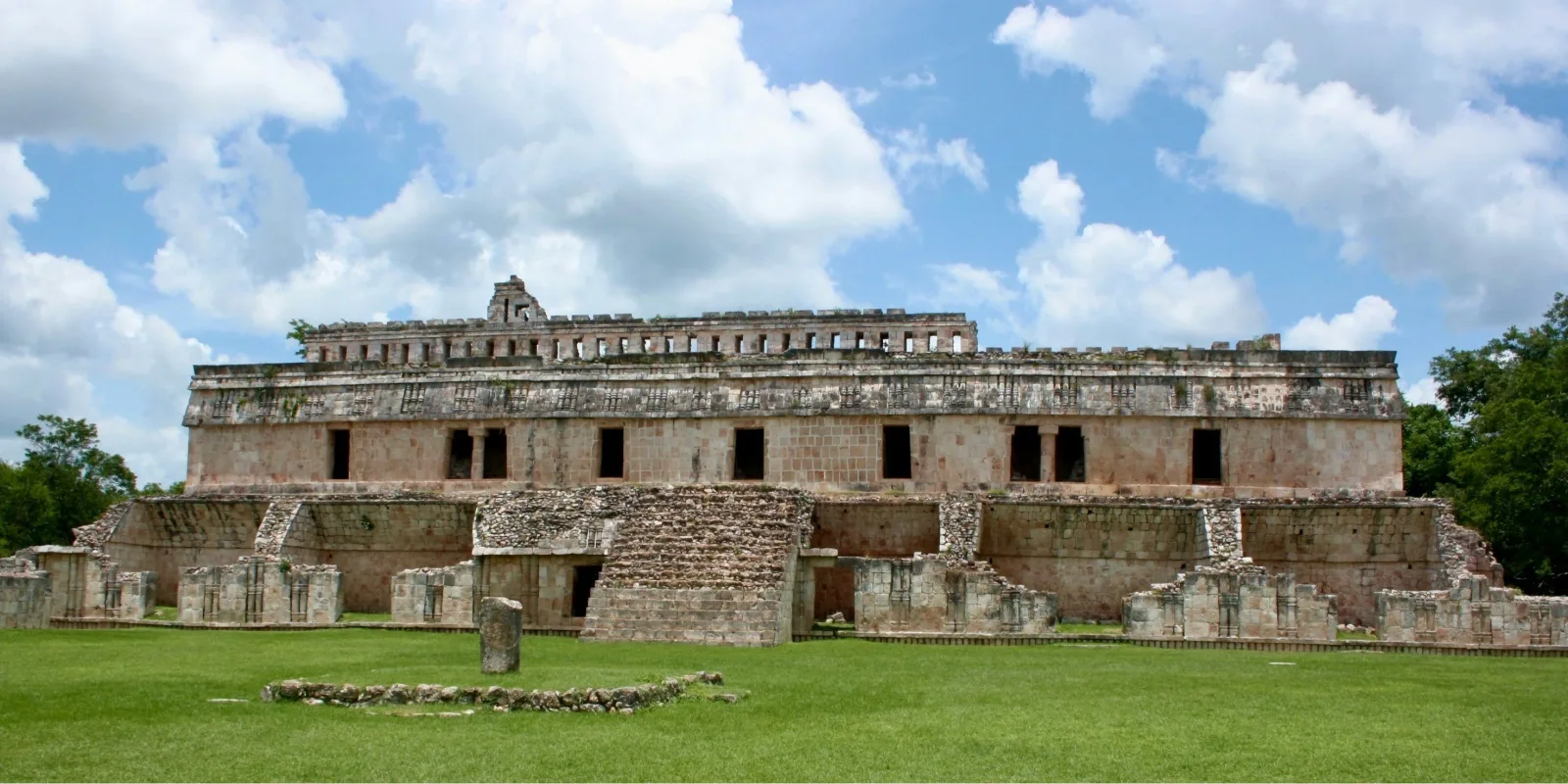 Wide-angle view of the Codz Poop Palace at Kabah, featuring intricate Puuc-style stonework and a raised ceremonial platform under a bright Yucatán sky.