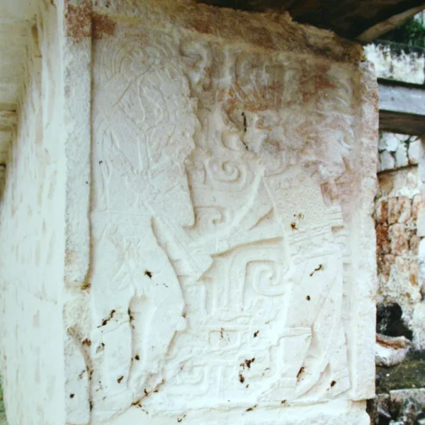 Close-up of a detailed Maya relief carving on a corner pillar at Kabah, showing stylized figures and swirling motifs in light-colored stone.