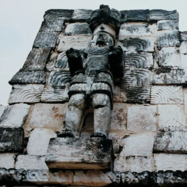 A detailed stone carving of a Maya warrior with feathered headdress on the façade of a palace at Kabah, part of the ancient Puuc ruins.