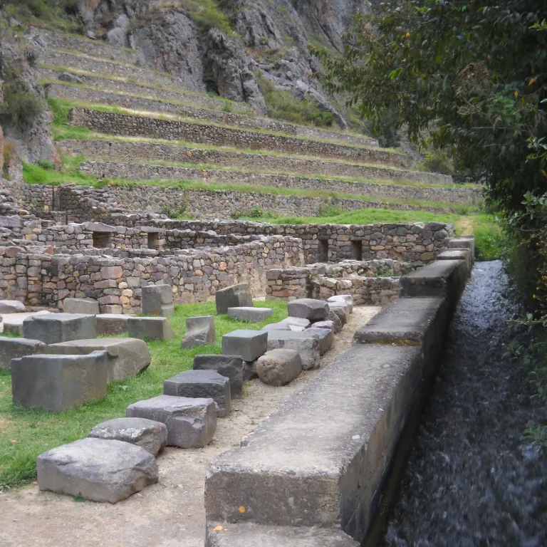 Stone canal running beside Inca ruins and agricultural terraces at Ollantaytambo, showcasing advanced water engineering.