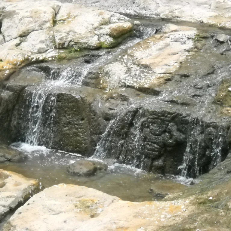 San Agustín stone carving partially submerged under a waterfall, featuring a sculpted face integrated into the natural rock