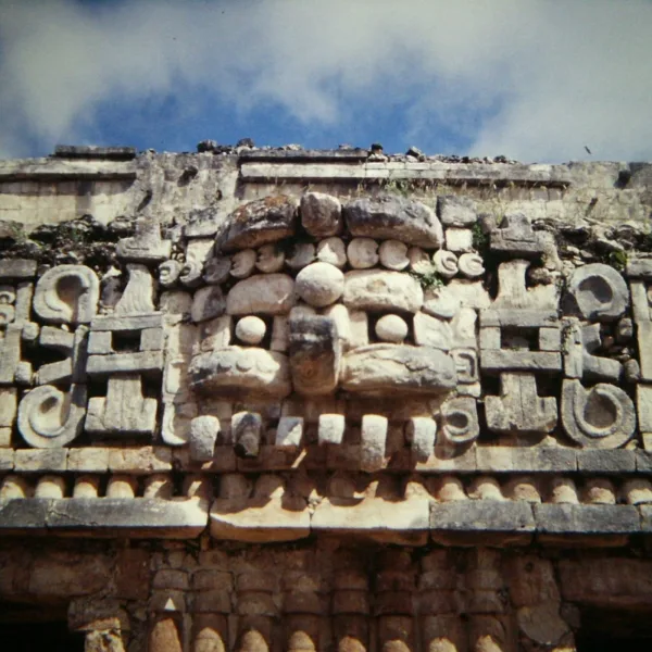 Close-up of a carved Chaac mask on a Puuc-style temple at Sayil, featuring intricate mosaic stonework and symbolic rain god iconography under a bright sky
