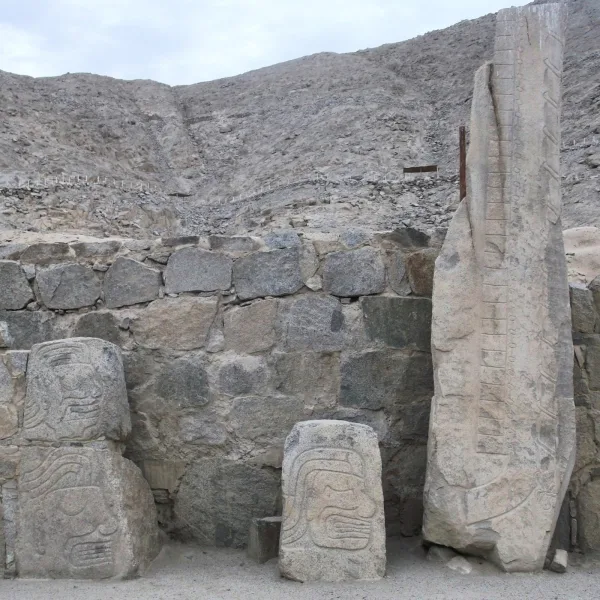 Tall engraved stone monoliths and carved faces at Cerro Sechín, backed by a rocky Andean hillside
