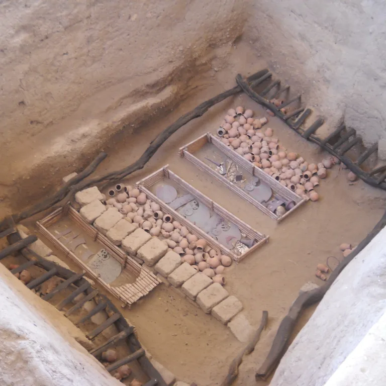 Overhead view of an excavated Moche tomb at Sipán showing rows of ceramic vessels, metal ornaments, and wooden support beams surrounding burial chambers.