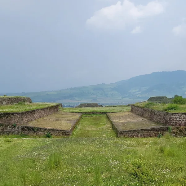 View down the center of Teotenango’s stone ballcourt, framed by low walls and backed by green hills under a misty afternoon sky