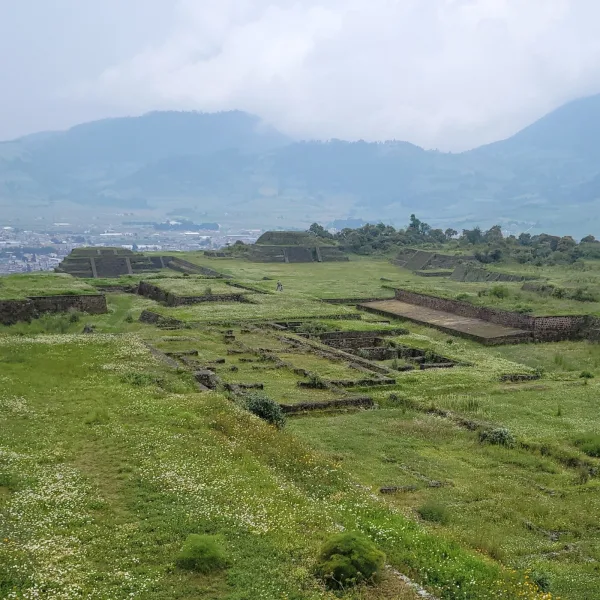 A wide view of Teotenango’s terraced ruins and residential platforms, with mountains and lowland cityscape visible in the distance under clouded skies