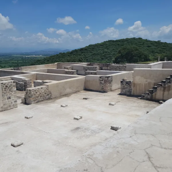 View of multi-room stone residential complex atop the acropolis at Xochicalco, with surrounding green hills in the background.