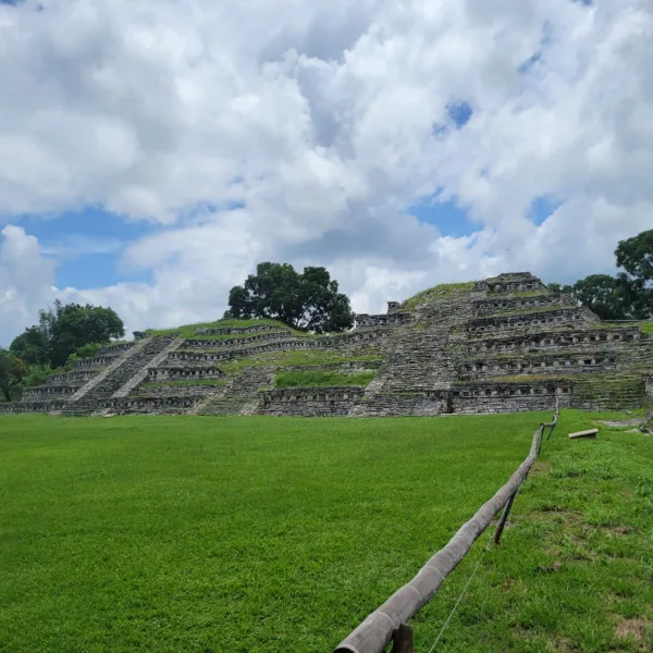 A broad green plaza at Yohualichan surrounded by low stone walls and stairways, with a jungle hill rising in the background.
