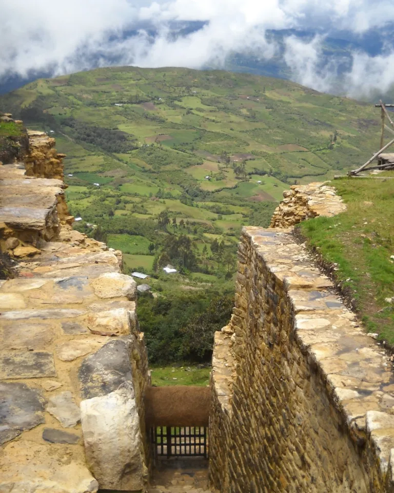 View from Kuélap ruins showing a narrow stone corridor leading down the mountain, with the green Utcubamba Valley visible through clouds in the background