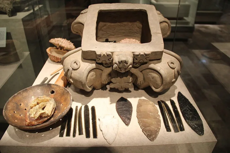 Zapotec ceramic offering box from Monte Albán with obsidian blades, flint tools, and ritual items on display in a museum collection.