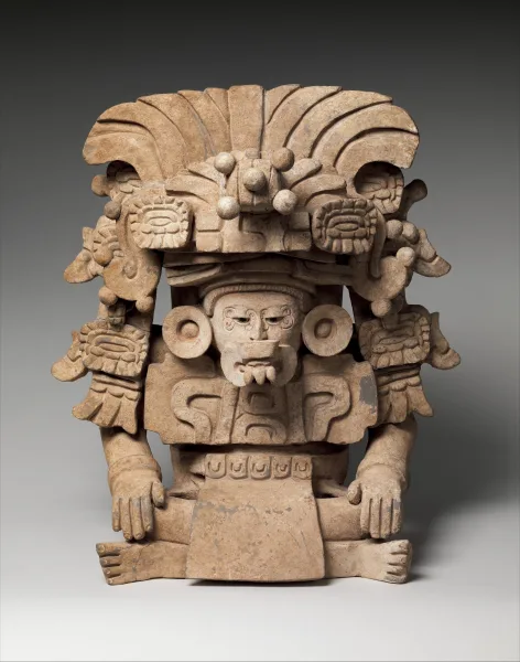 Classic Period Zapotec ceramic funerary effigy urn (350–600 CE) representing a priest or ancestor figure known as 7 Ñ (Seven Scroll/Knot). The seated figure emerges from a cylindrical vessel, wearing an elaborate buccal mask, earflares, and a complex feathered headdress, typical of Monte Albán tombs.