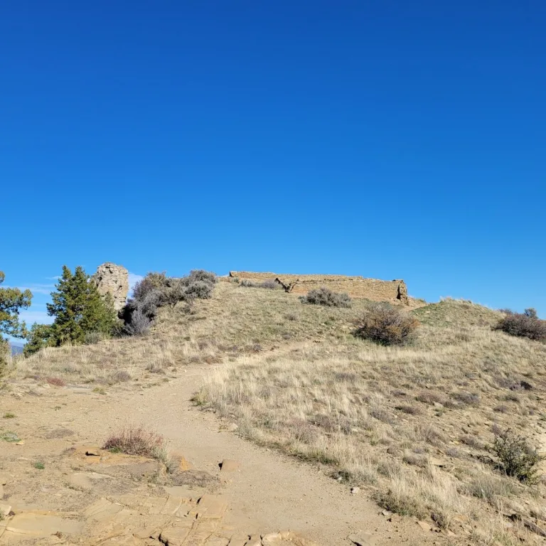 Dirt trail winding up a grassy, sunlit mesa towards the stone ruins of the Ancestral Puebloan Great House Complex against a bright blue sky.