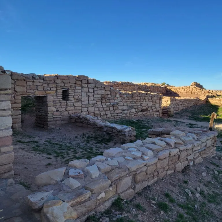Panoramic view of the Lowry Pueblo site, showing the low excavated stone foundations and surrounding mesa landscape under a bright sky.