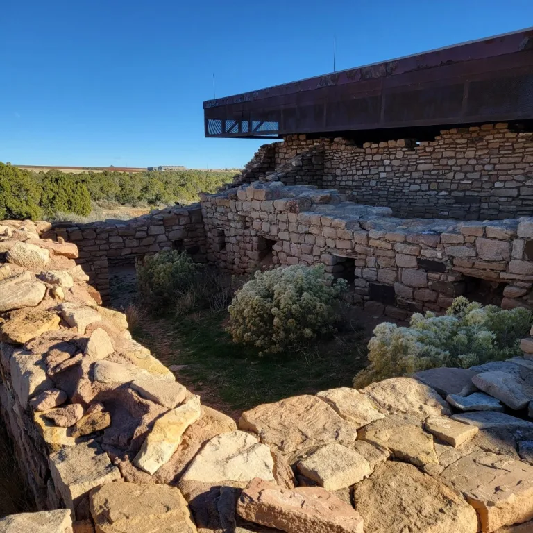 Ground-level view of a standing segment of the rectangular sandstone masonry wall of the main pueblo room block under a clear sky.