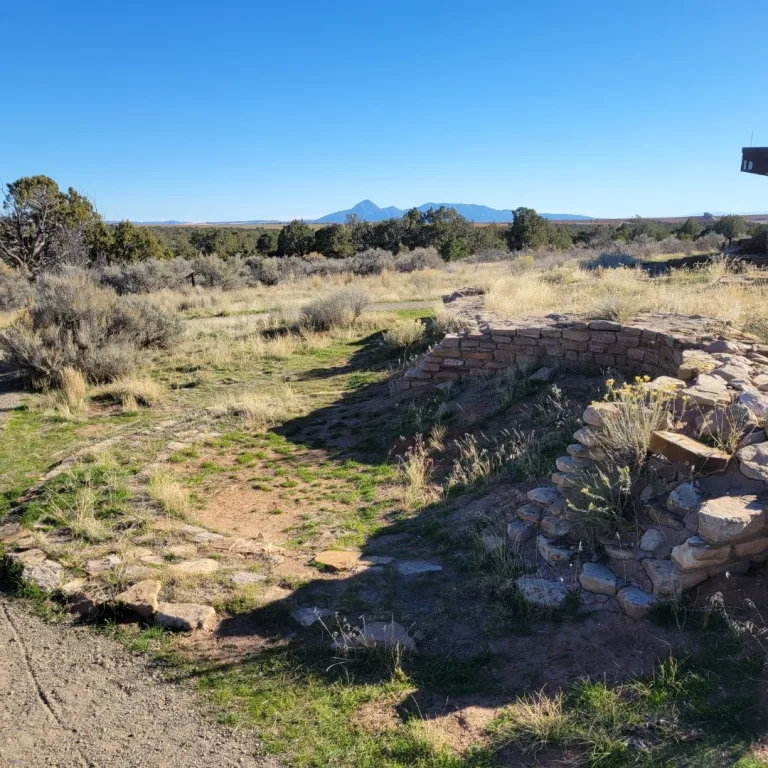 View of the Lowry Pueblo ruins on the mesa, with the excavated foundations in the foreground and the distinct profile of Ute Mountain (Ute Peak) rising prominently in the background.
