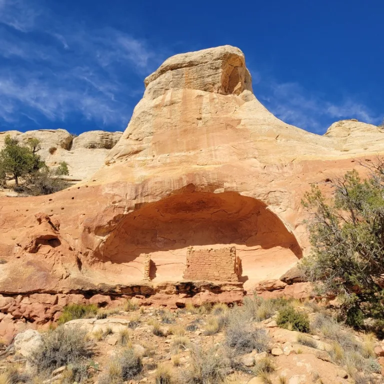 A small, stone room block known as the Saddlehorn Pueblo, built into a deep alcove beneath a distinctive saddle-shaped capstone rock formation.