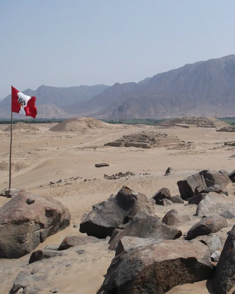 Wide view overlooking the archaeological ruins of Caral in the Supe Valley of Peru, with stone platform mounds and plazas spread across a desert landscape and a Peruvian flag in the foreground.