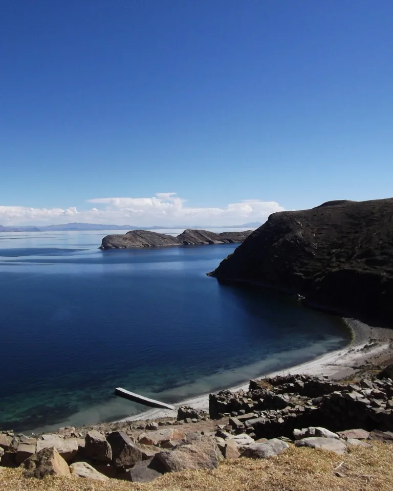 View of the rocky shoreline and calm blue waters of Lake Titicaca on Isla del Sol, with stone terraces and hills rising above the lake.