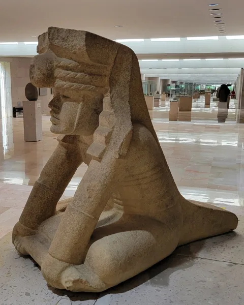 “Olmec stone sculpture of a kneeling figure with forward-placed arms and a tall ridged headdress displayed in the MAX, the Museo de Antropología de Xalapa.