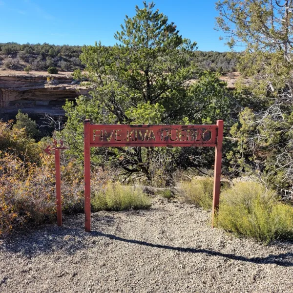 Red metal trail sign reading Five Kiva Pueblo standing beside a gravel path with pinyon and juniper trees and canyon cliffs in the background in Utah.