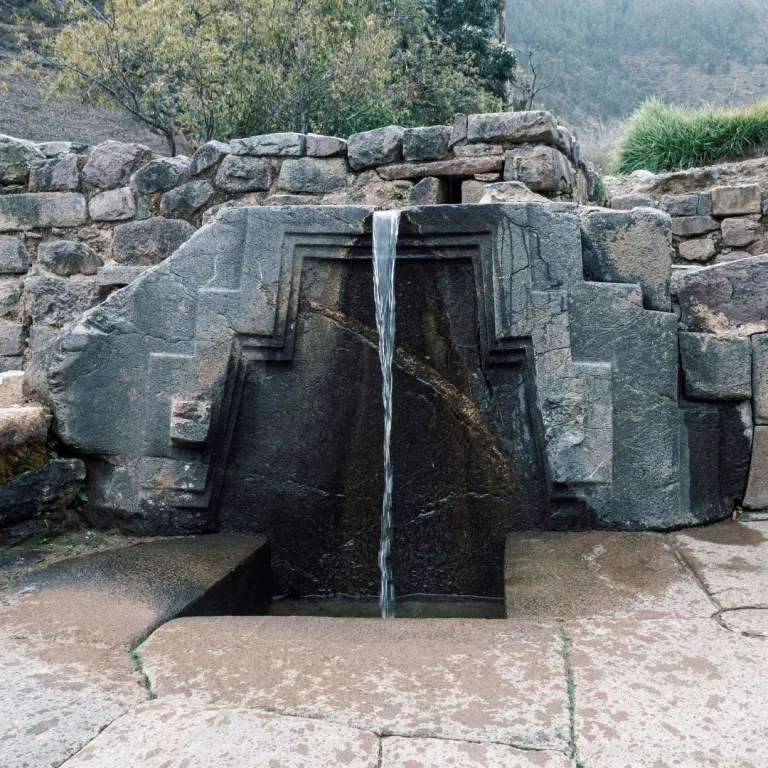 Stone fountain known as Baño de la Ñusta at Ollantaytambo in Peru showing a carved trapezoidal niche with water flowing into a rectangular basin surrounded by Inca stone masonry.