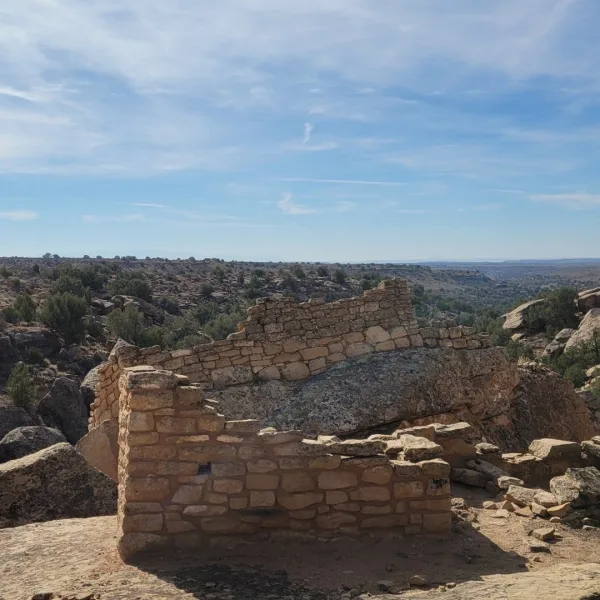 Low masonry walls at the Holly Group in Hovenweep National Monument, built around and against a large sandstone boulder, with pinyon-juniper canyon landscape in the background.