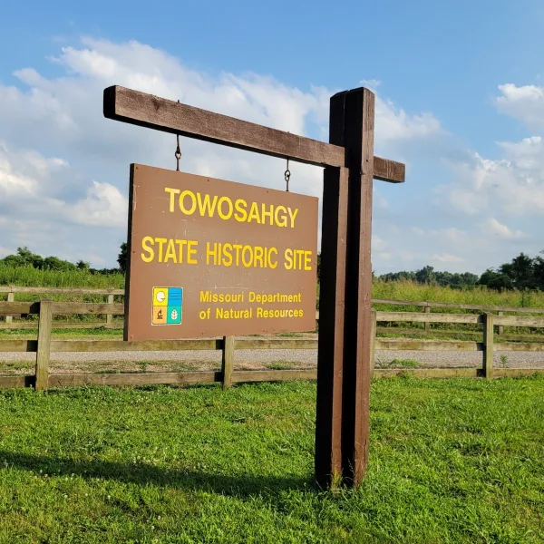 Entrance sign for Towosahgy State Historic Site in Missouri, with a wooden fence, grass, and open sky behind it.