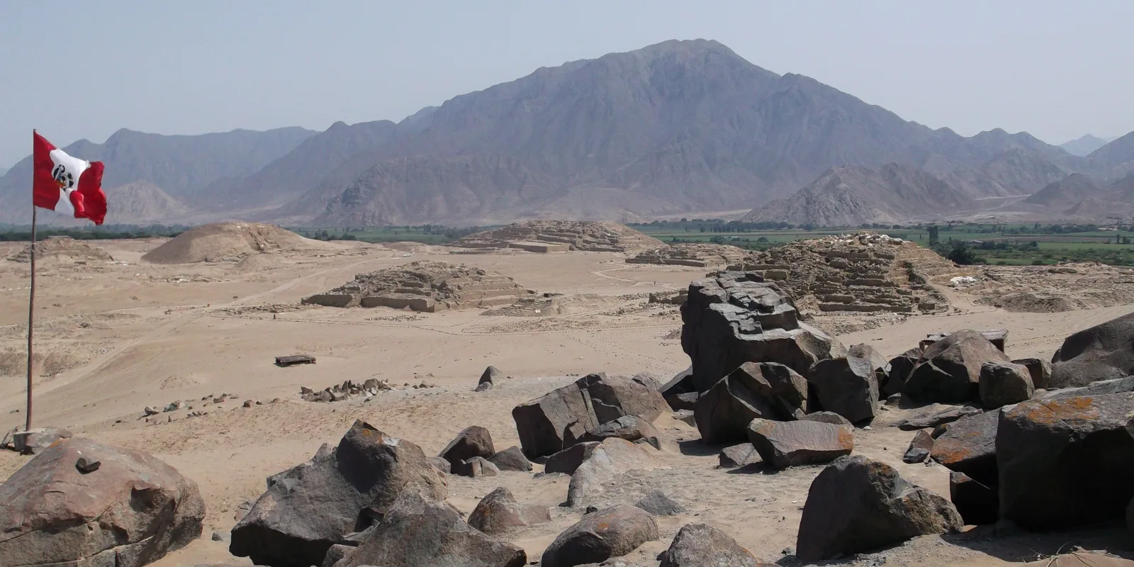 Ancient pyramid structures at the Sacred City of Caral in Peru, with a Peruvian flag, desert terrain, foreground rocks, and Supe Valley mountains in the background.