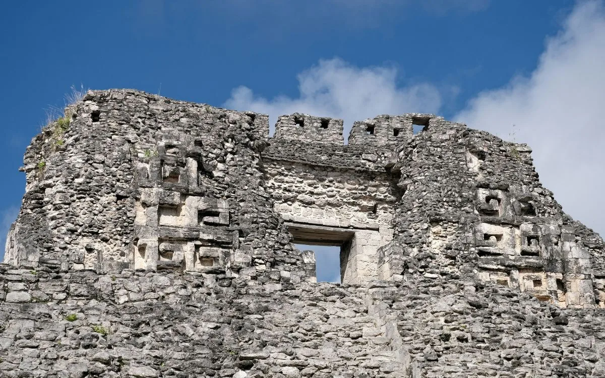 Upper east facade of Structure X at Becán in Campeche, Mexico, showing a central doorway flanked by surviving Chenes-style zoomorphic stone mosaic mask elements.