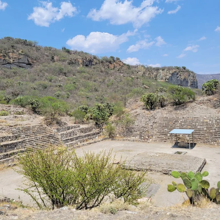 A stone courtyard at the Zapotec site of Yagul in Oaxaca, Mexico, with stairways, a low platform, cacti, and rocky hills behind it.