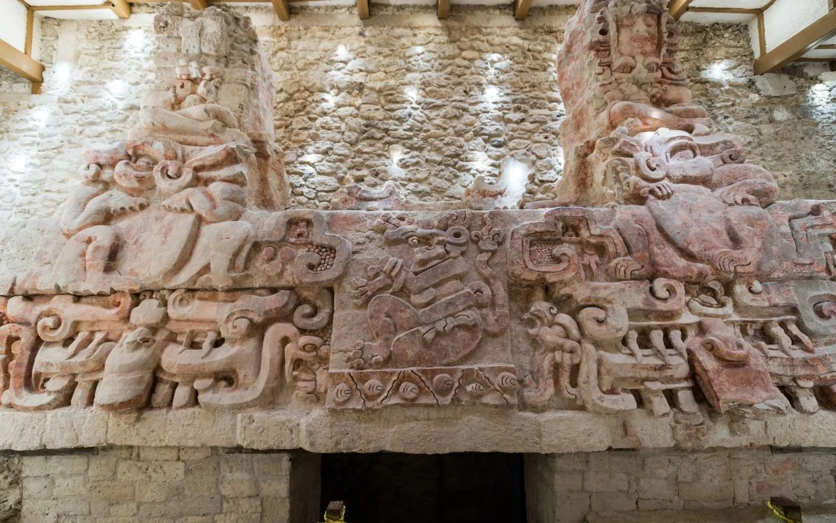 Polychrome stucco frieze on Substructure I at Balamkú in Campeche, Mexico, showing elaborate Maya sculpted faces, zoomorphic figures, curling decorative motifs, and surviving red pigment above a stone entrance.