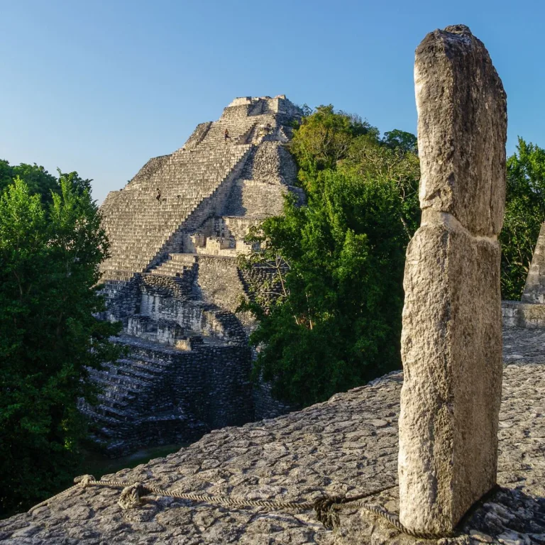 Structure IX at Becán rises beyond the trees, with its steep staircase visible behind a stone pillar on Structure VIII.
