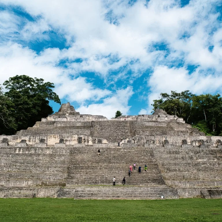 Front view of Caana at Caracol in Cayo District, Belize, showing its monumental central staircase, broad stone terraces, palace doorways, and visitors climbing beneath a cloudy blue sky.