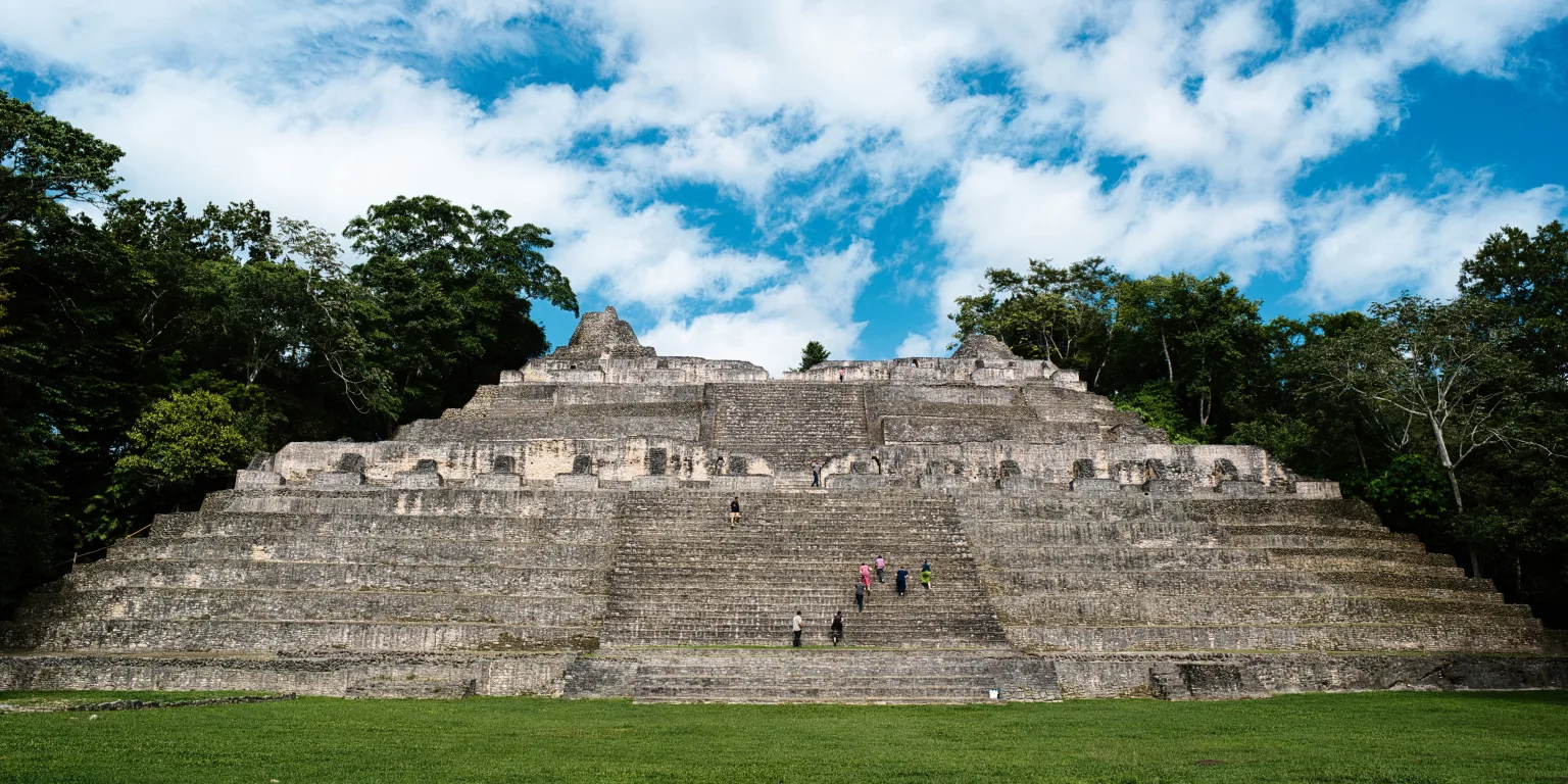 Wide Front view of Caana at Caracol in Cayo District, Belize, showing its monumental central staircase, broad stone terraces, palace doorways, and visitors climbing beneath a cloudy blue sky.
