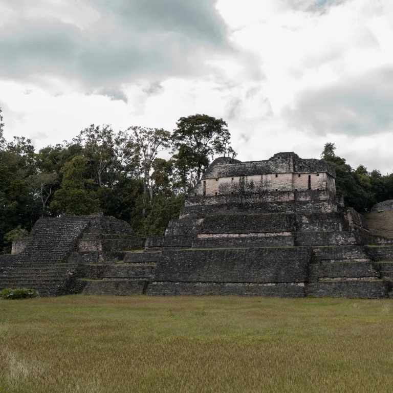 Rear view of Structure A6 at Caracol in Cayo District, Belize, showing its standing summit temple walls, narrow openings, stepped stone base, and adjoining ruins beneath an overcast sky.
