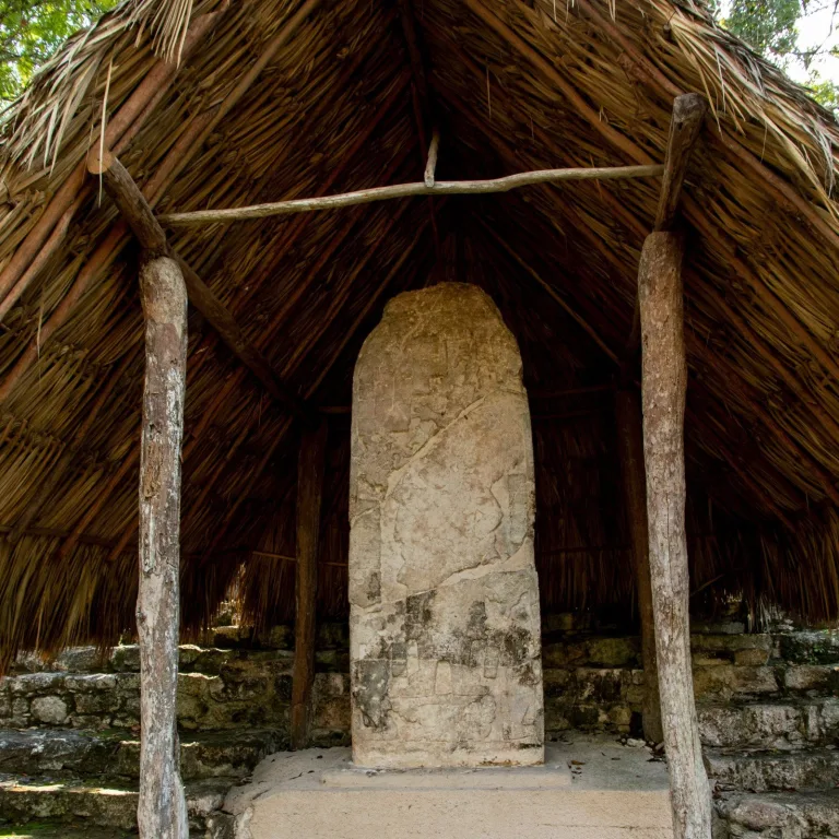 Weathered Stela 20 at Cobá, Mexico, showing a carved Maya ruler, faint hieroglyphs, and diagonal fractures beneath a palm-thatched shelter.