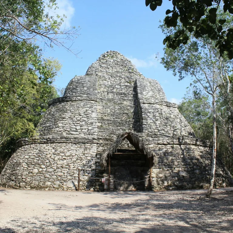 Xaibé at Cobá in Quintana Roo, Mexico, showing four rounded stone tiers, a steep central stepped feature, and a small protective thatched shelter at its base, surrounded by forest.