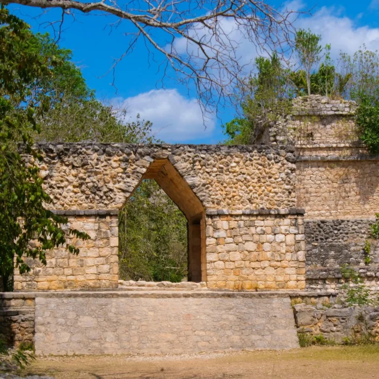 The pointed limestone entrance arch at Ek Balam in Yucatán, Mexico, stands on a low platform with trees visible through its opening.