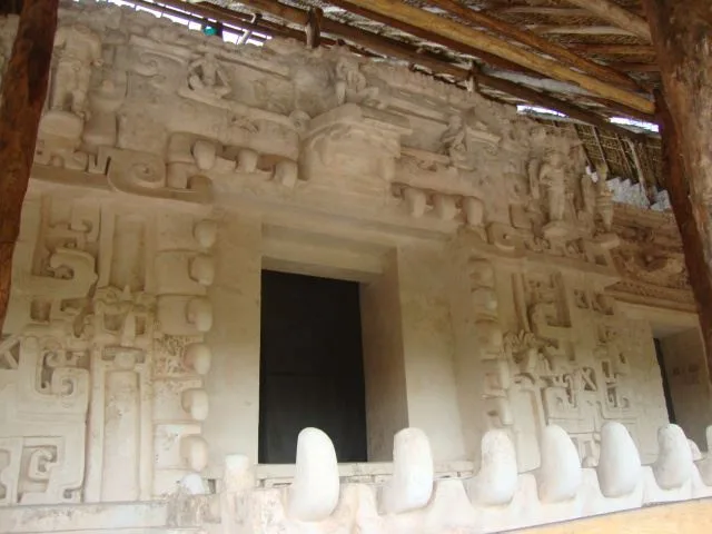 Sak Xok Naah stucco facade in the Acropolis at Ek Balam, Yucatán, Mexico, showing sculpted figures, intricate reliefs, and projecting teeth around a dark doorway beneath a protective roof.