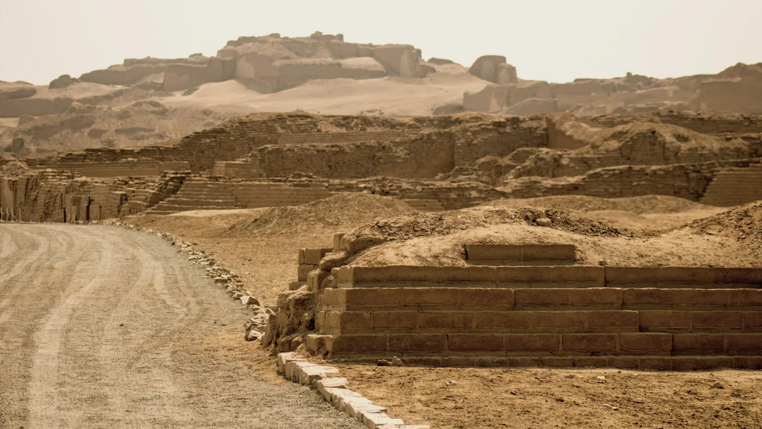 Adobe and stone ruins at Pachacamac near Lima, Peru, showing stepped masonry walls, a winding dirt road, and monumental terraced structures rising across the desert landscape.