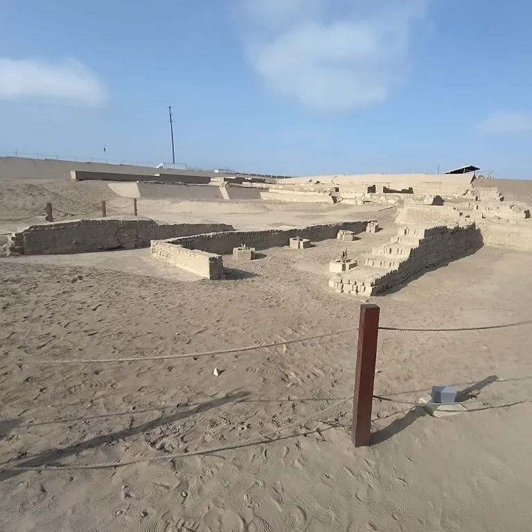 Low adobe walls form rectangular enclosures at Pachacamac, with small raised bases inside a roped-off excavation area.